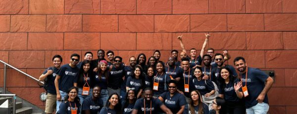 A large group of people in matching Wharton School t-shirts posing in front of a building sign that reads 