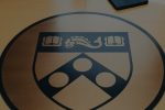 A table with the University of Pennsylvania emblem on it, featuring a coat of arms with elements like books and a dolphin. A backpack and a laptop are also on the table.