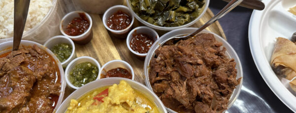 An assortment of traditional dishes on a wooden board, including rice, various meats in sauces, leafy greens, and small cups of sauce or condiments.