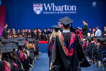 A graduation ceremony with graduates in caps and gowns walking toward a stage. The background shows a sign for Wharton, University of Pennsylvania.