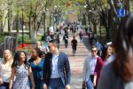 A group of people walking along a tree-lined pathway in a sunny park or campus setting. The atmosphere is lively and social.