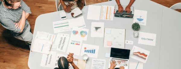 A small group of people in a meeting, gathered around a table with laptops, charts, and documents labeled with terms like 