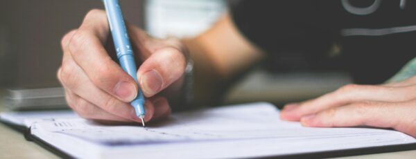 A person writing in a notebook with a blue pen, focusing on the hands and pen on a lined page.